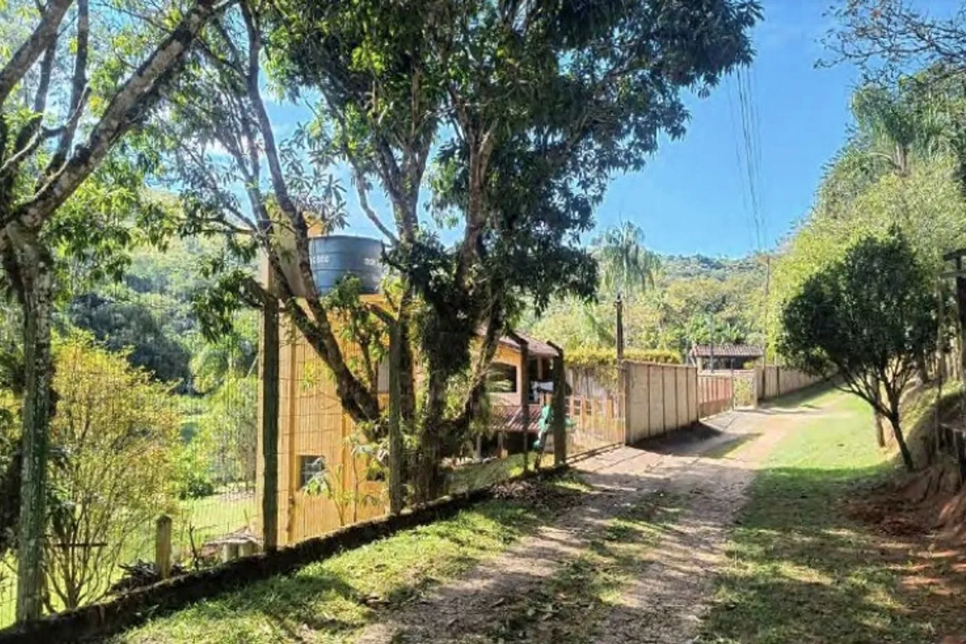 Acesso de terra ao imóvel, com porteira de madeira e construções ao fundo.